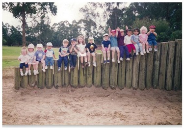 Young children smiling and posing while sitting in a row on log stumps at a park