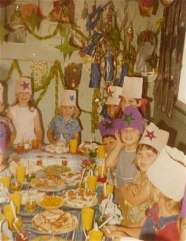Young children wearing party hats, sitting around a table of food at the Family Day Care Christmas party