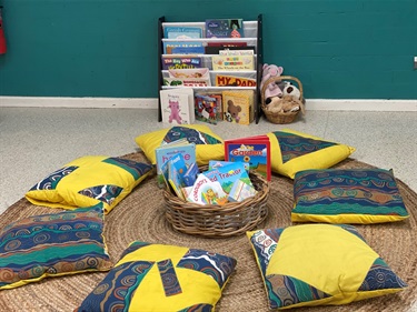 A basket of children's books sitting on a circular brown mat with yellow cushions placed around it in the children's reading area