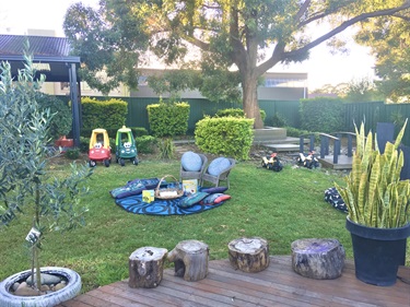 A mat and chairs with a basket of books, and different toy ride on cars located on the grass area of the children's outdoor area