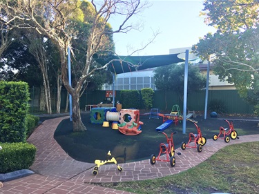 Colourful playground equipment on dark green rubber flooring under the shade, besides a line of tricycles parked on the path